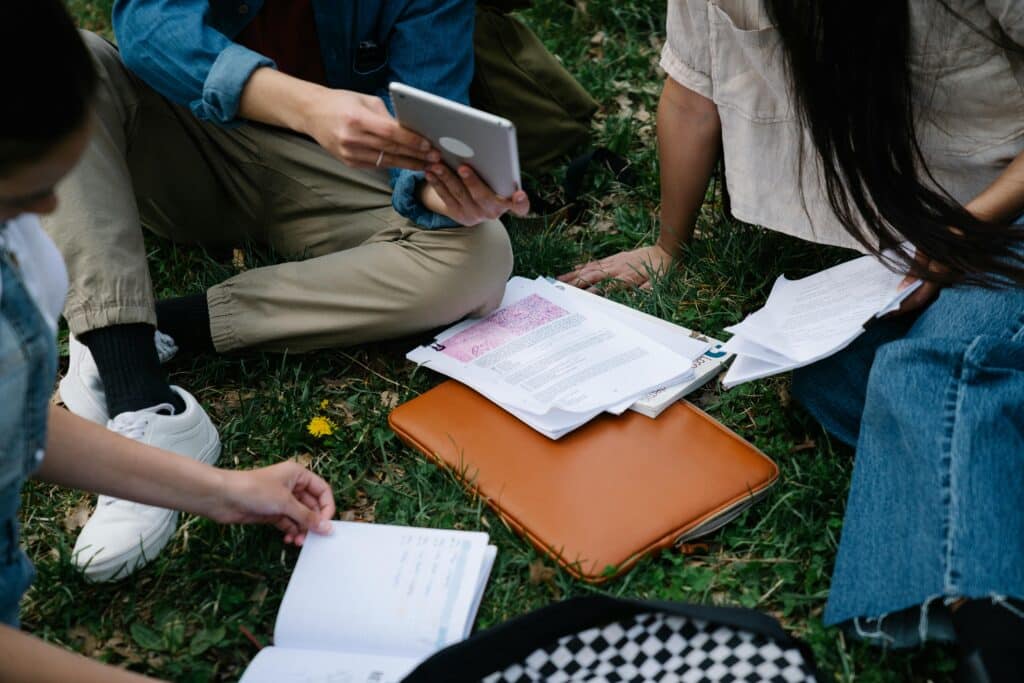 Group of people sitting on grass outdoors, studying with notebooks, papers, and a tablet nearby.